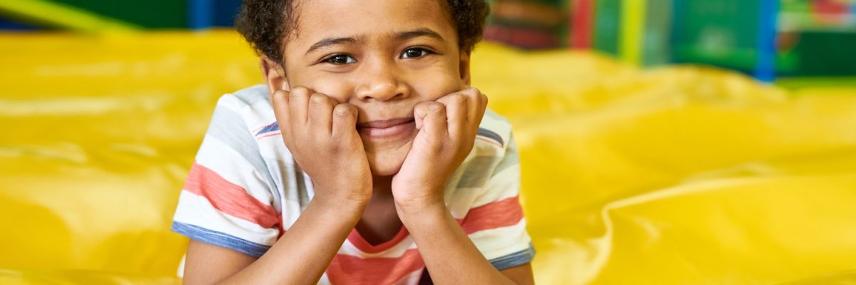 Colorful portrait of cute African-American boy smiling happily at camera while having fun in bouncy castle of children play center, copy space